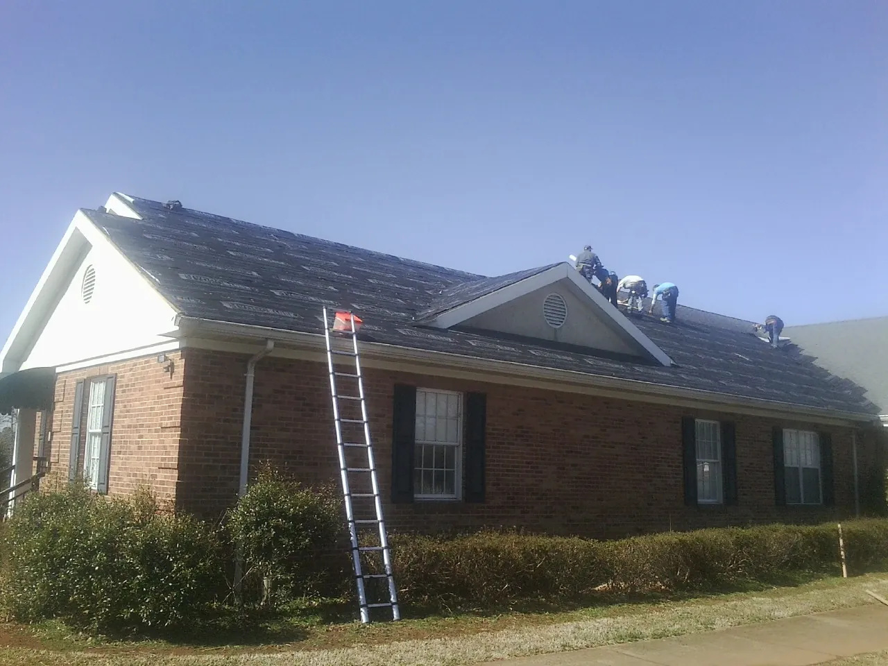 Crew tearing shingles off a brick ranch roof with a ladder set — Emergency Roof Repair work by our crew in Camp Hill