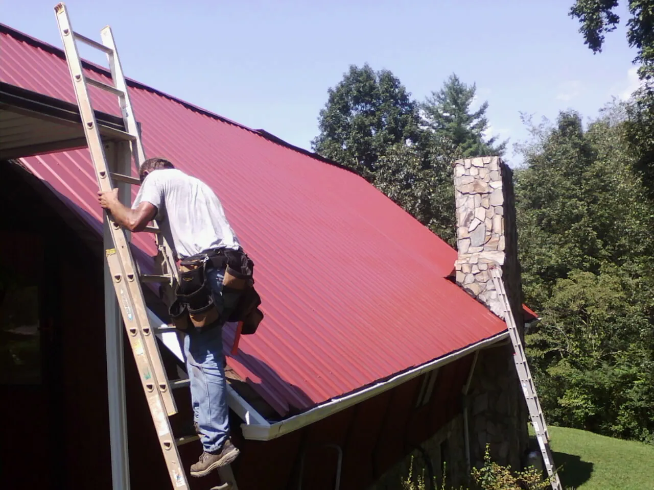 Roofer on a ladder beside a red metal roof and stone chimney — Soffit & Fascia Repair by Roofing Brothers in Camp Hill